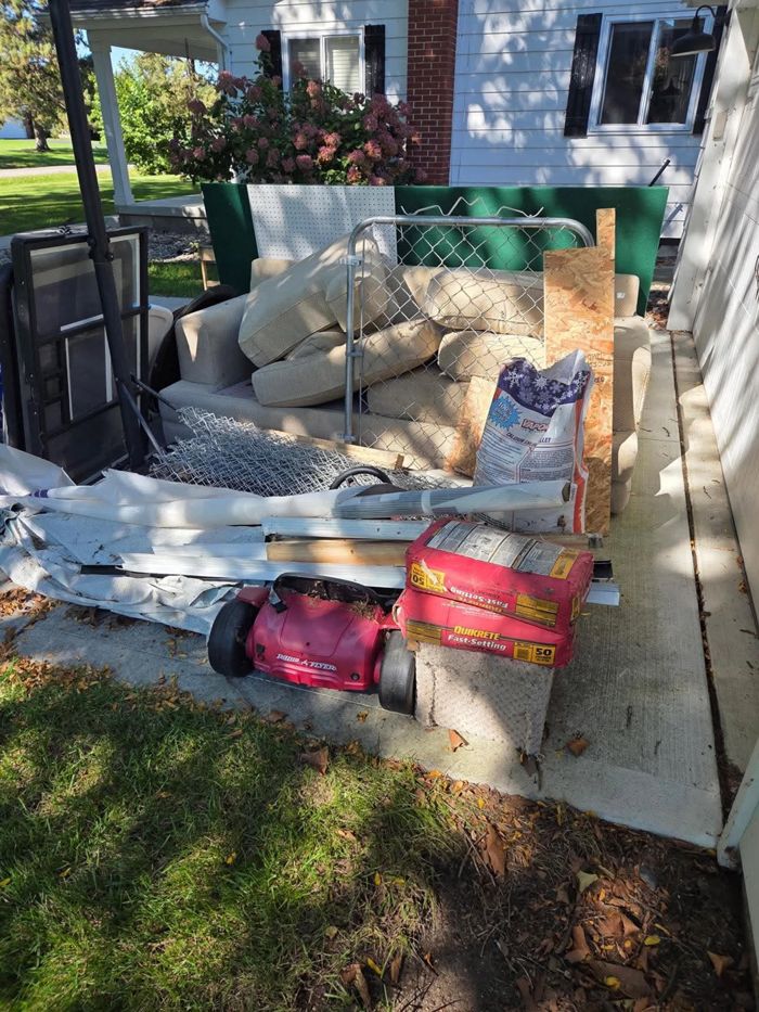 A pile of junk beside a house: an old couch, chain-link fence and gate, plywood, bags of cement and a red toy ride-on car